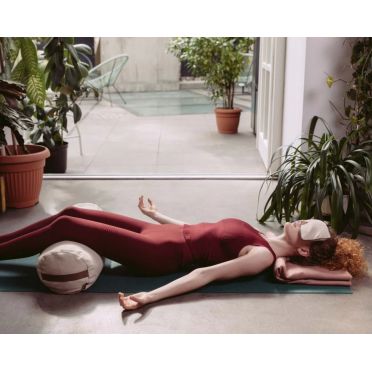 Kobieta w bordowym stroju do jogi leży na macie w pozycji relaksacyjnej z lawendową poduszką na oczy do relaksacji i poduszką pod kolanami, w tle rośliny i szklane drzwi prowadzące na patio.